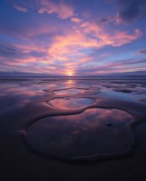A photograph captures a sunset sky mirrored in tidal pools on a beach, rendered in a palette of cool blues, greens, and purpl