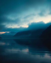 A glassy fjord reflects a sunset, enveloped in a soft cinematic haze. The scene is dominated by rich deep teal tones, part of