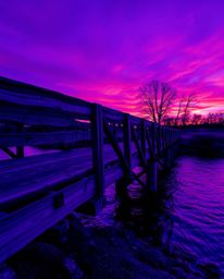 An old wooden bridge over a river at sunset is reimagined in striking, deep ultraviolet hues. The scene is characterized by v