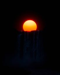 An intense jet black sun setting behind a dramatic waterfall, depicted with pure black, deep shadows, and vibrant light strea