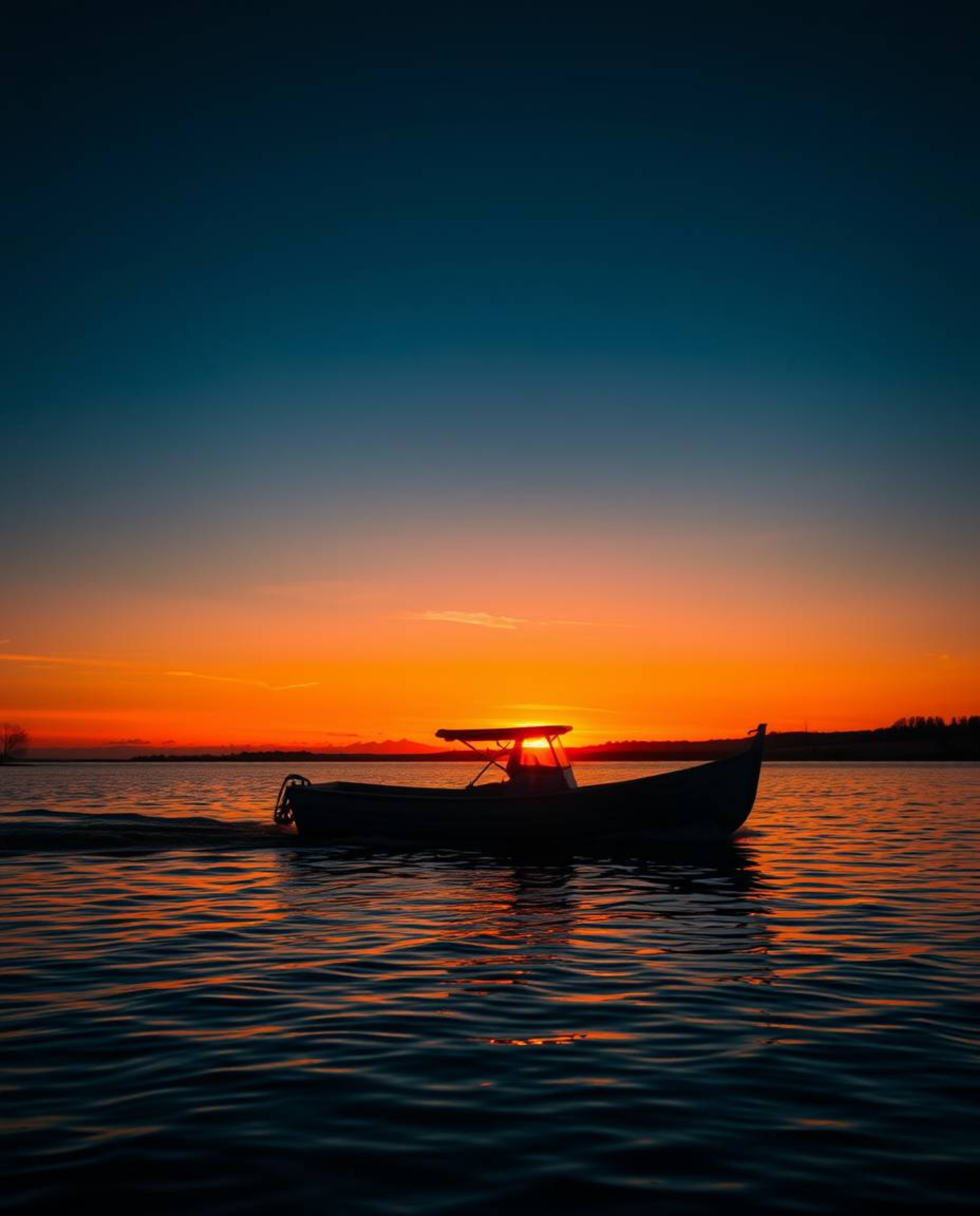 High-contrast, low-key lighting accentuates a boat drifting on a lake during sunset. The intentional use of deep shadows crea