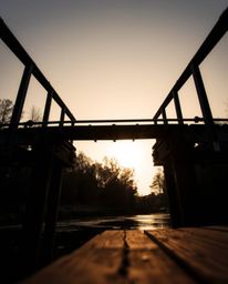 A low-angle perspective of an old wooden bridge over a river at sunset. The image is rendered in an intense jet black with de