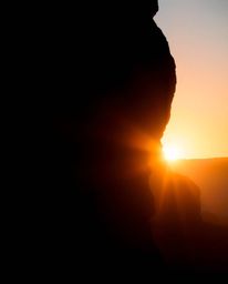 A silhouetted image, created using dramatic backlighting, depicts a sunset illuminating sandstone cliffs in a canyon. The art