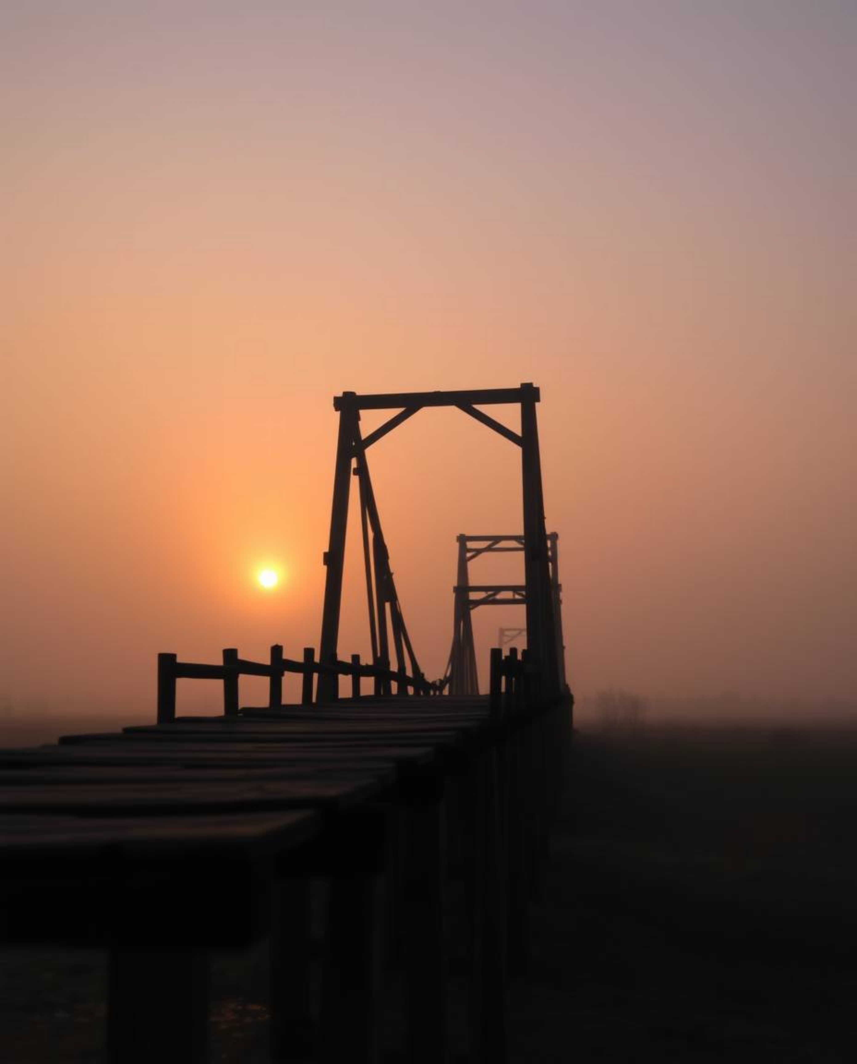 This cinematic photograph captures an old wooden bridge over a river at sunset, enveloped in a soft haze. The scene is distin