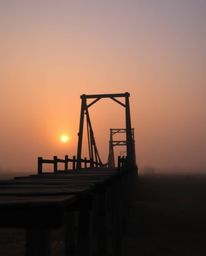 This cinematic photograph captures an old wooden bridge over a river at sunset, enveloped in a soft haze. The scene is distin