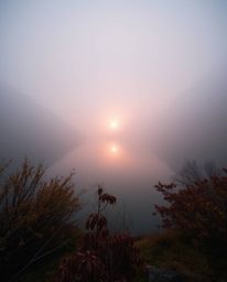 The reflection of a sunset in a glassy fjord is depicted amidst thick foggy surroundings. This artwork is characterized by it