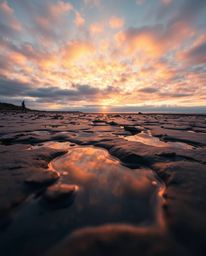 A low-angle perspective photograph captures a striking sunset sky as it reflects in the tidal pools of a beach. The scene is 