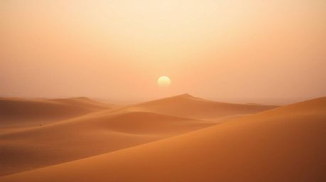 Experience a soft, cinematic sunset at the edge of a mesmerizing desert landscape, adorned with rolling golden dunes. The haz