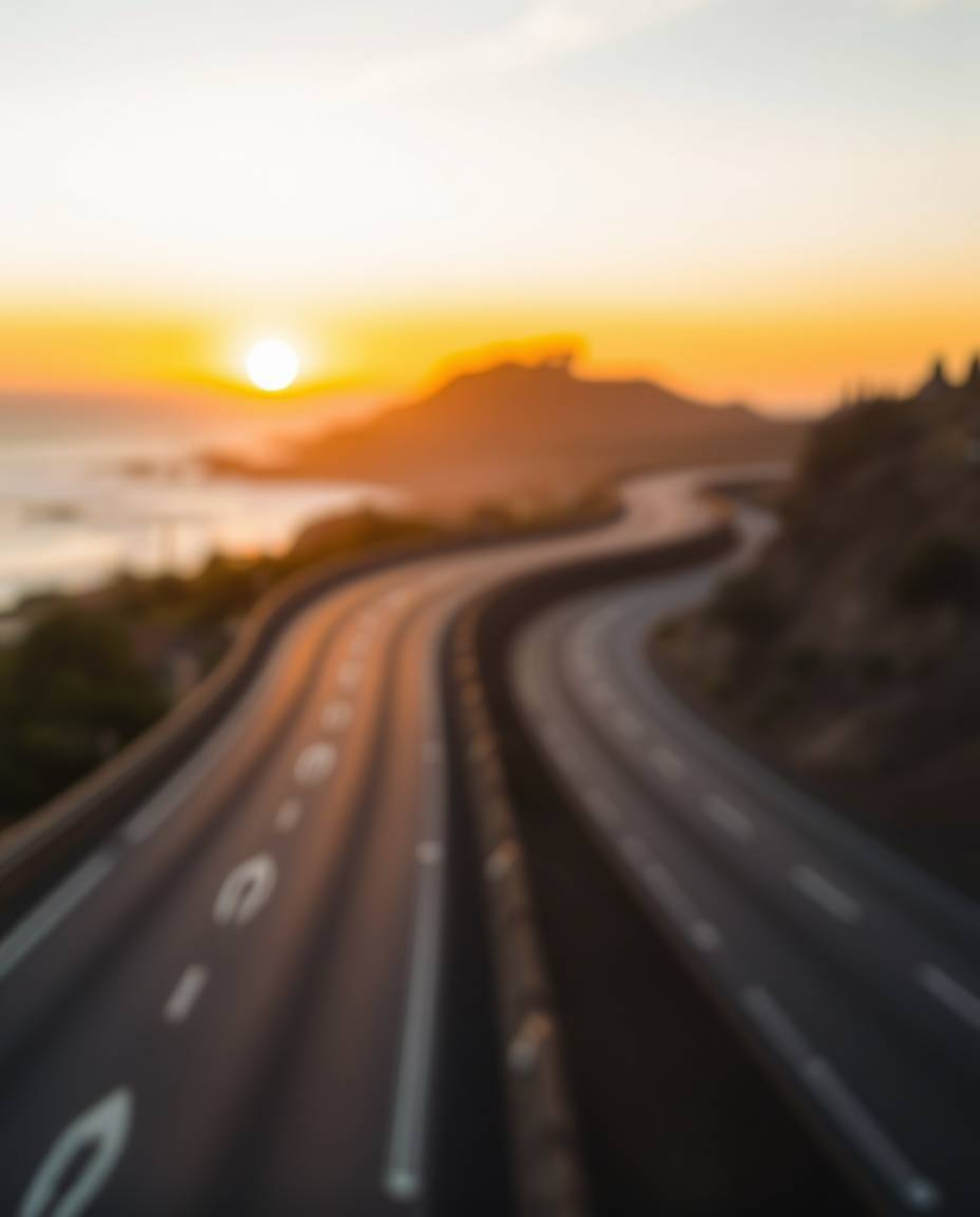 This photograph captures a winding coastal highway at sunset, featuring a prominent blurred bokeh effect that creates a soft,