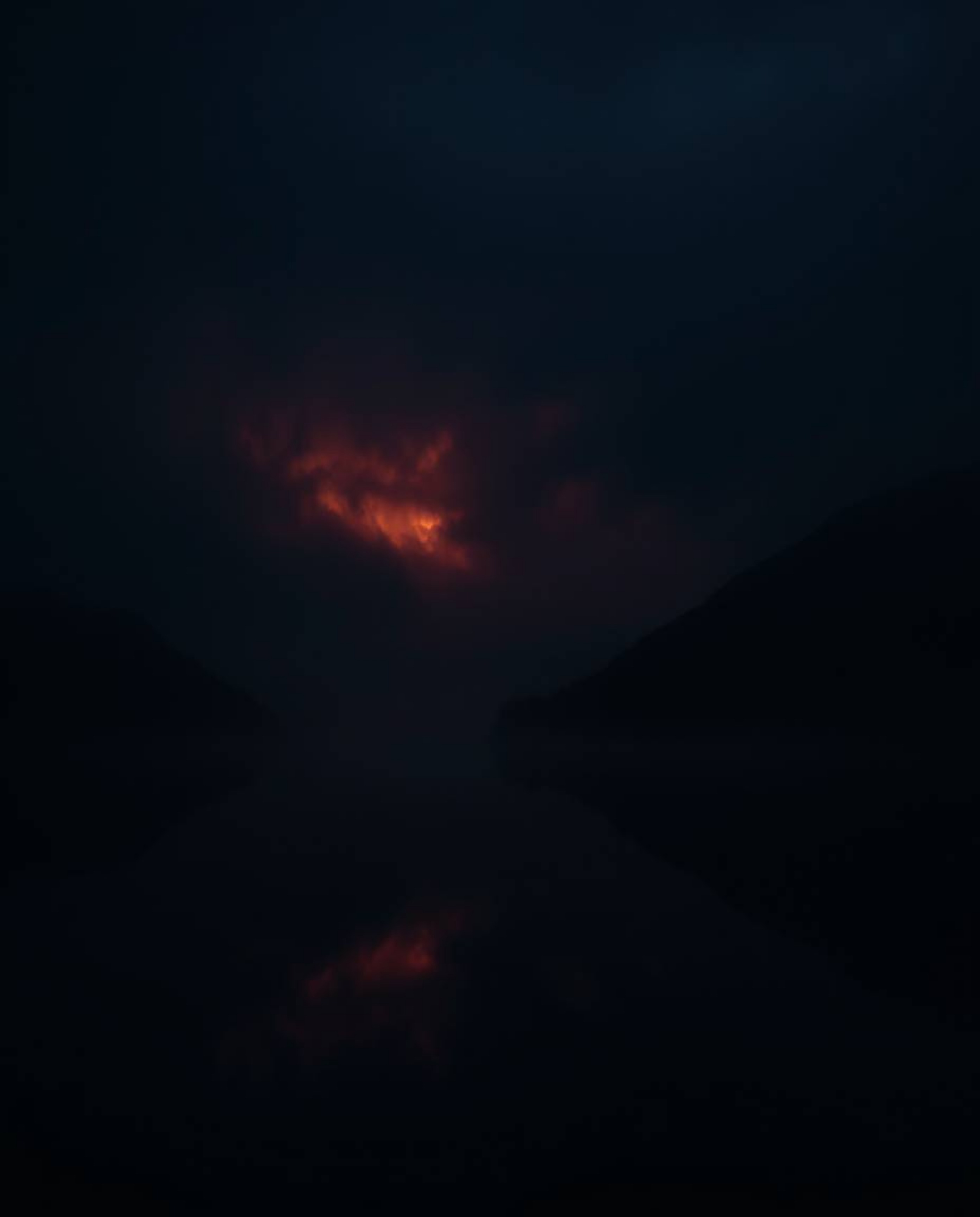 A photograph depicting the reflections of fiery clouds in a mountain lake, enveloped in intense jet black and thick fog. The