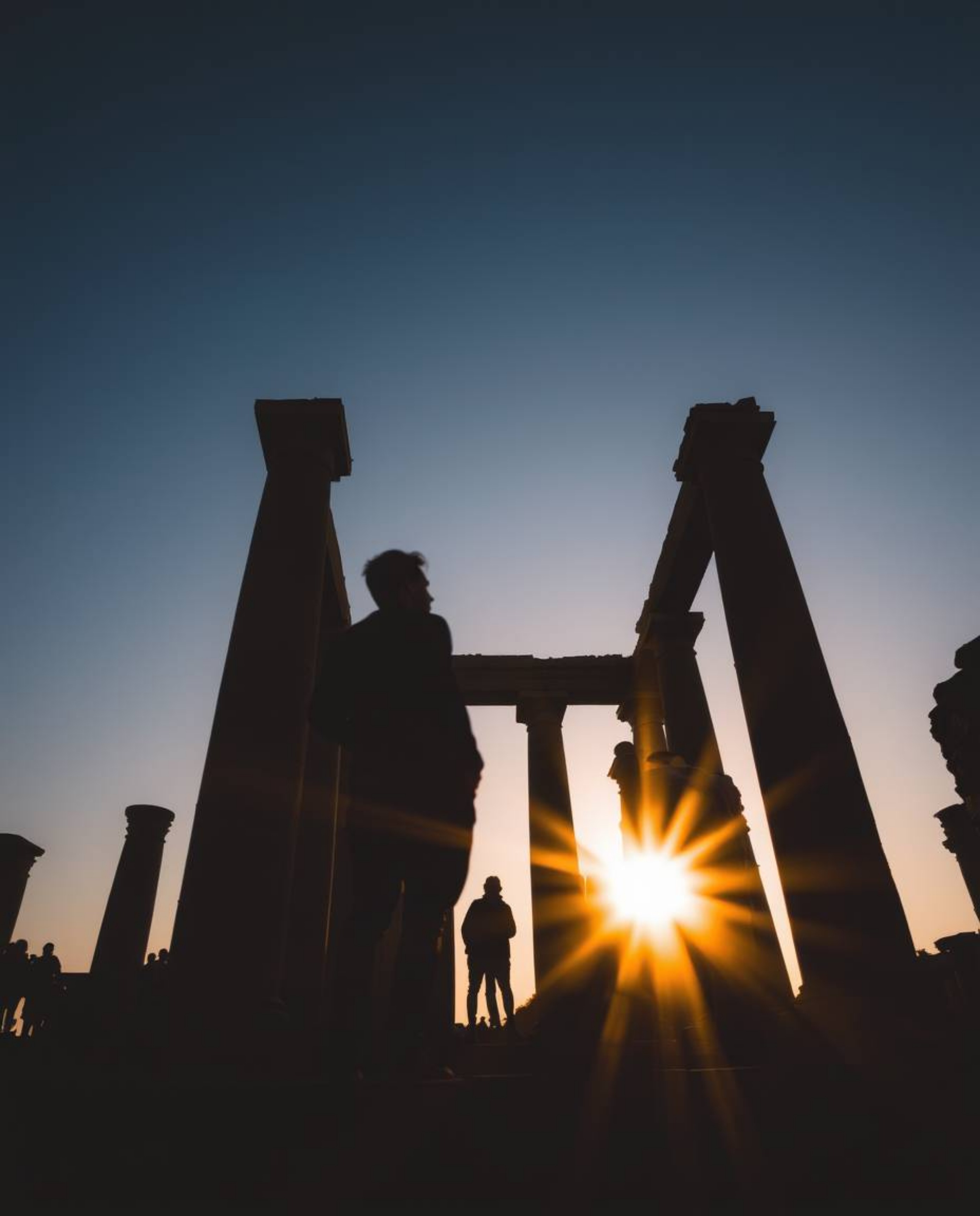 Silhouetted figures are captured in a long-exposure low-angle photograph of ancient stone ruins at sunset. From a perspective