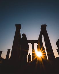 Silhouetted figures are captured in a long-exposure low-angle photograph of ancient stone ruins at sunset. From a perspective