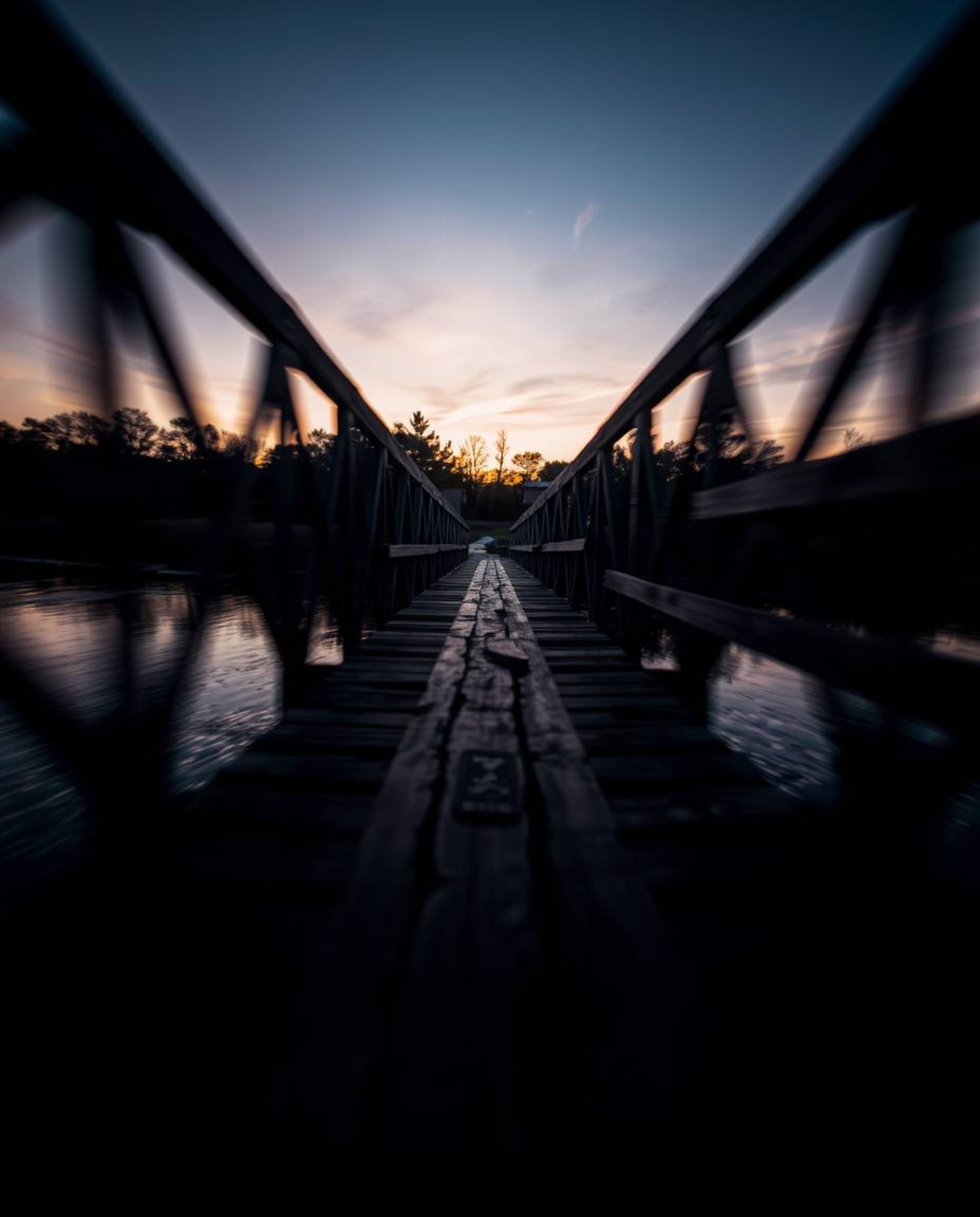 A high-contrast, low-key photograph of an old wooden bridge over a river at sunset. This striking image showcases blurred lin