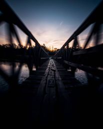 A high-contrast, low-key photograph of an old wooden bridge over a river at sunset. This striking image showcases blurred lin