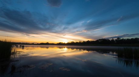 A captivating image of a serene pond reflecting a vibrant sunset. Cool-toned blues, greens, and purples dominate the sky's re