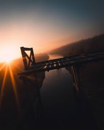 This moody photograph captures an old wooden bridge over a river during sunset from a dramatic high-angle perspective. The el