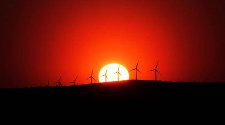 Capture the unsettling beauty of a sunset behind a row of wind turbines on a hill. This photograph utilizes dark tones and a 