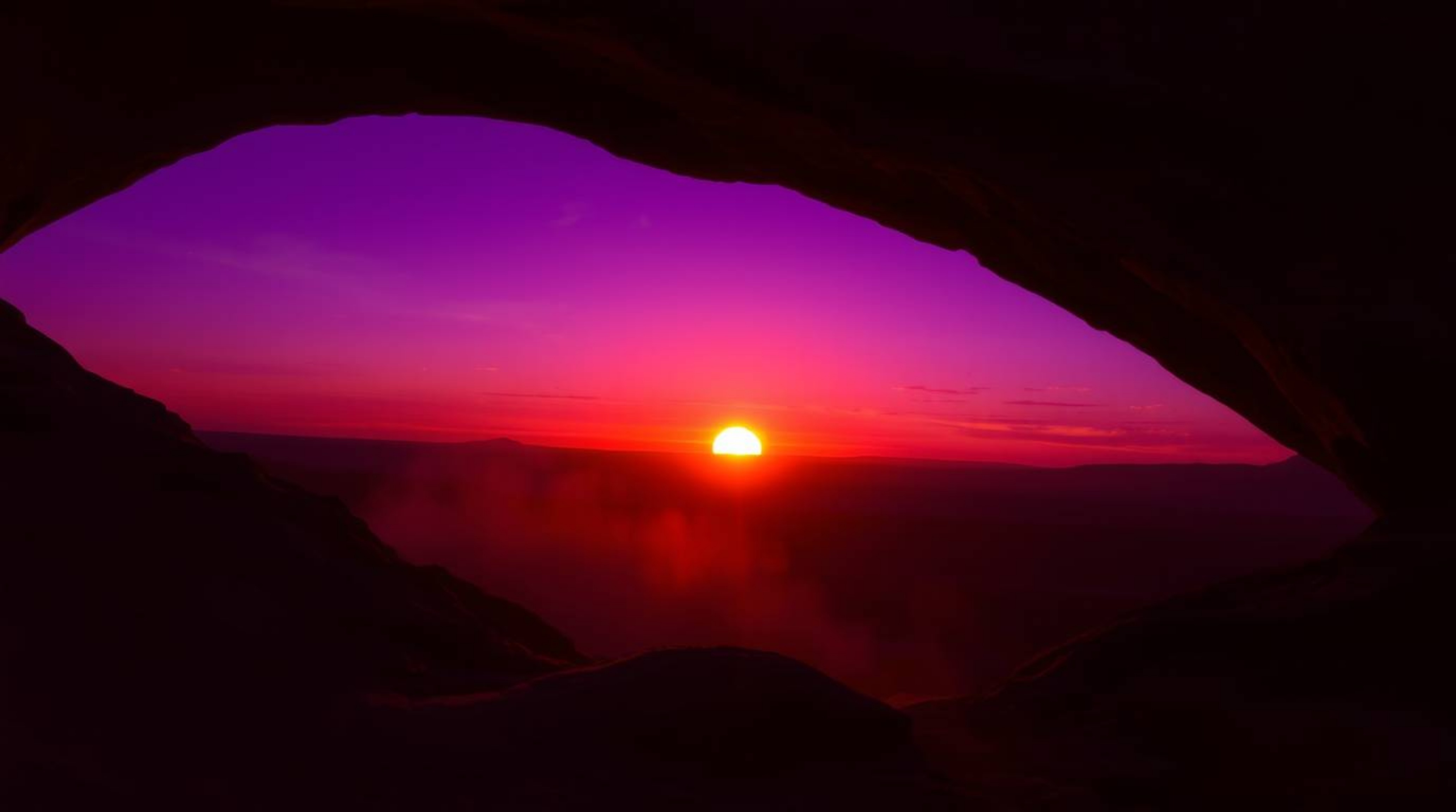 Witness the breathtaking spectacle of a desert sunset framed by a natural rock arch, painted in intense ultraviolet and purpl