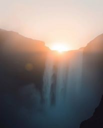 A soft, dreamy atmosphere with hazy soft light characterizes this scene of a sun setting behind a dramatic waterfall, creatin