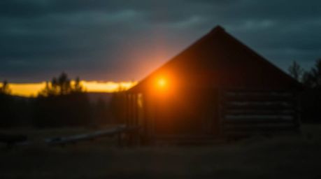 A strong, hard light creates a moody atmosphere in this image of an old wooden cabin at sunset, featuring a blurred bokeh eff