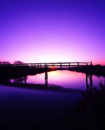 An image depicting an old wooden bridge over a river at sunset. The scene is dominated by deep ultraviolet hues, creating a g