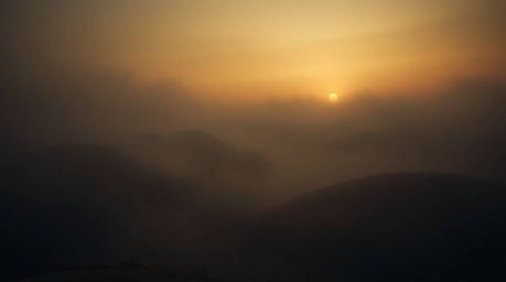 Capture the unsettling atmosphere of fog-covered hills bathed in the warm glow of a sunset. This image showcases volumetric l