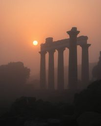 Soft cinematic haze envelops ancient stone ruins at sunset, creating a dreamy atmosphere. The scene is defined by its soft li