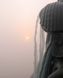A close-up shot of a bronze sculpture depicting a waterfall at sunset. Thick fog obscures the background, creating a low visi