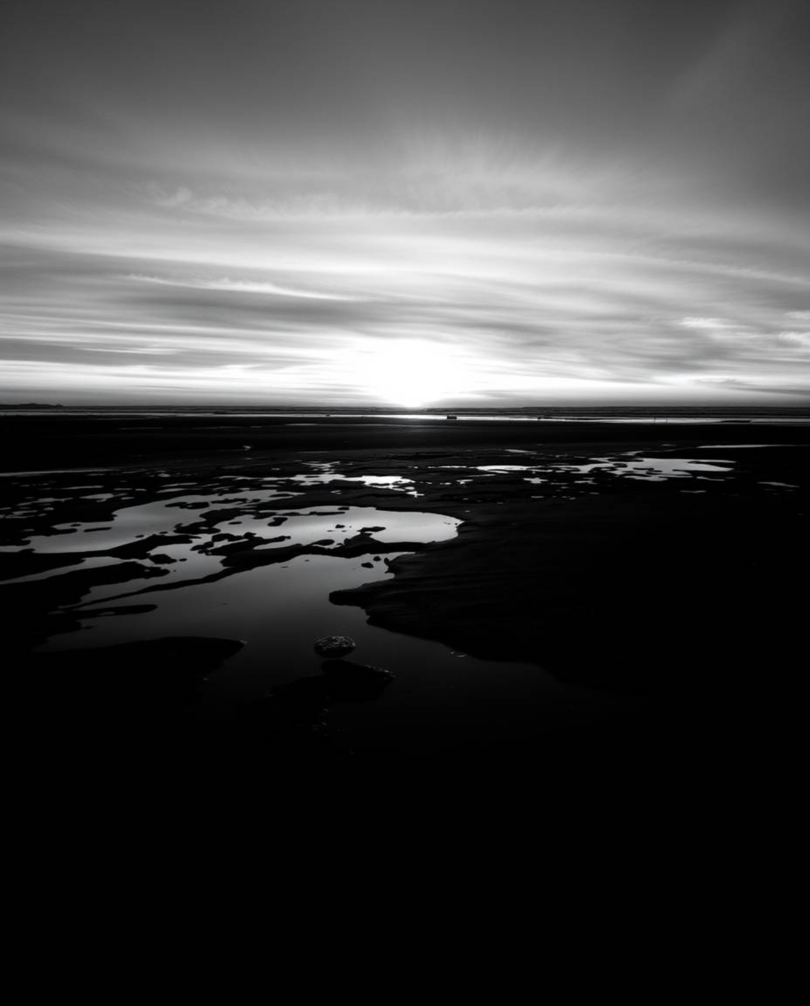 A classic black and white eerie photograph captures a sunset sky as it's mirrored in the tidal pools on a beach. The image cr
