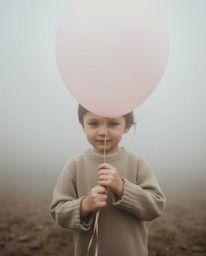 A captivating image of a child with fair skin gently holding a pastel-colored balloon amidst a thick fog. The muted browns an