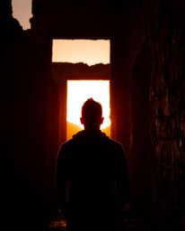 A silhouetted figure is backlit by a warm sunset that casts long shadows through ancient stone ruins. The scene emphasizes a 