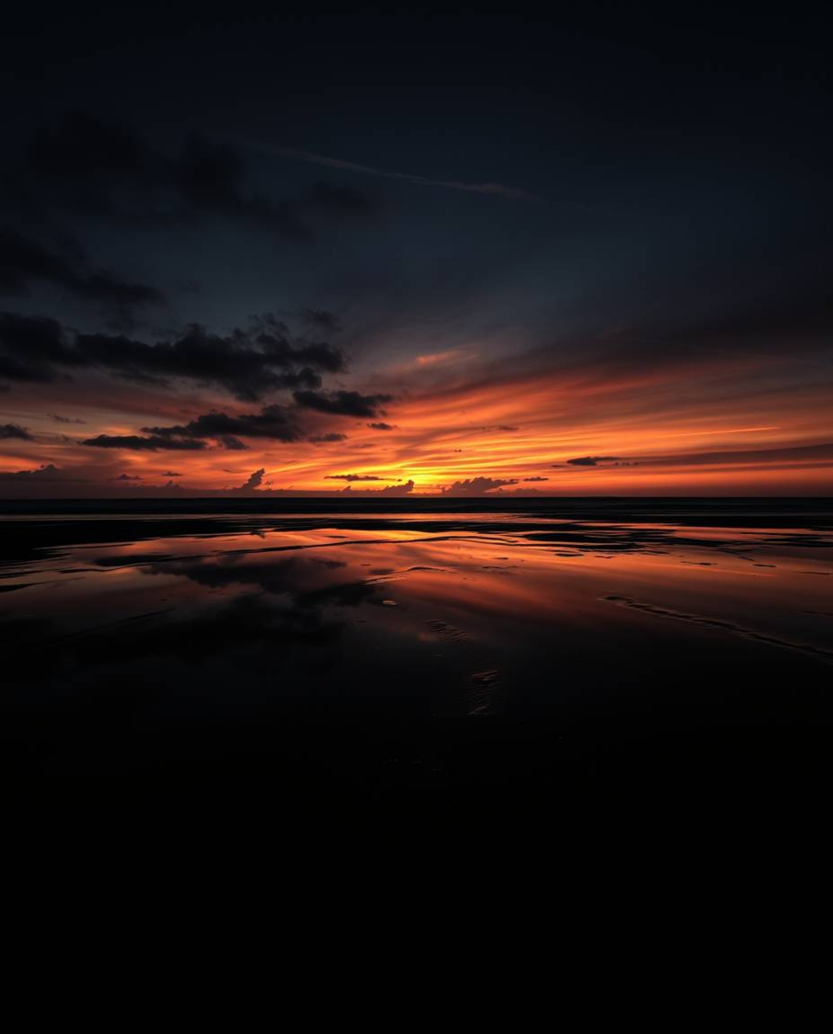 A dramatic chiaroscuro image depicts a sunset sky mirrored in the tidal pools of a beach. This powerful composition is define
