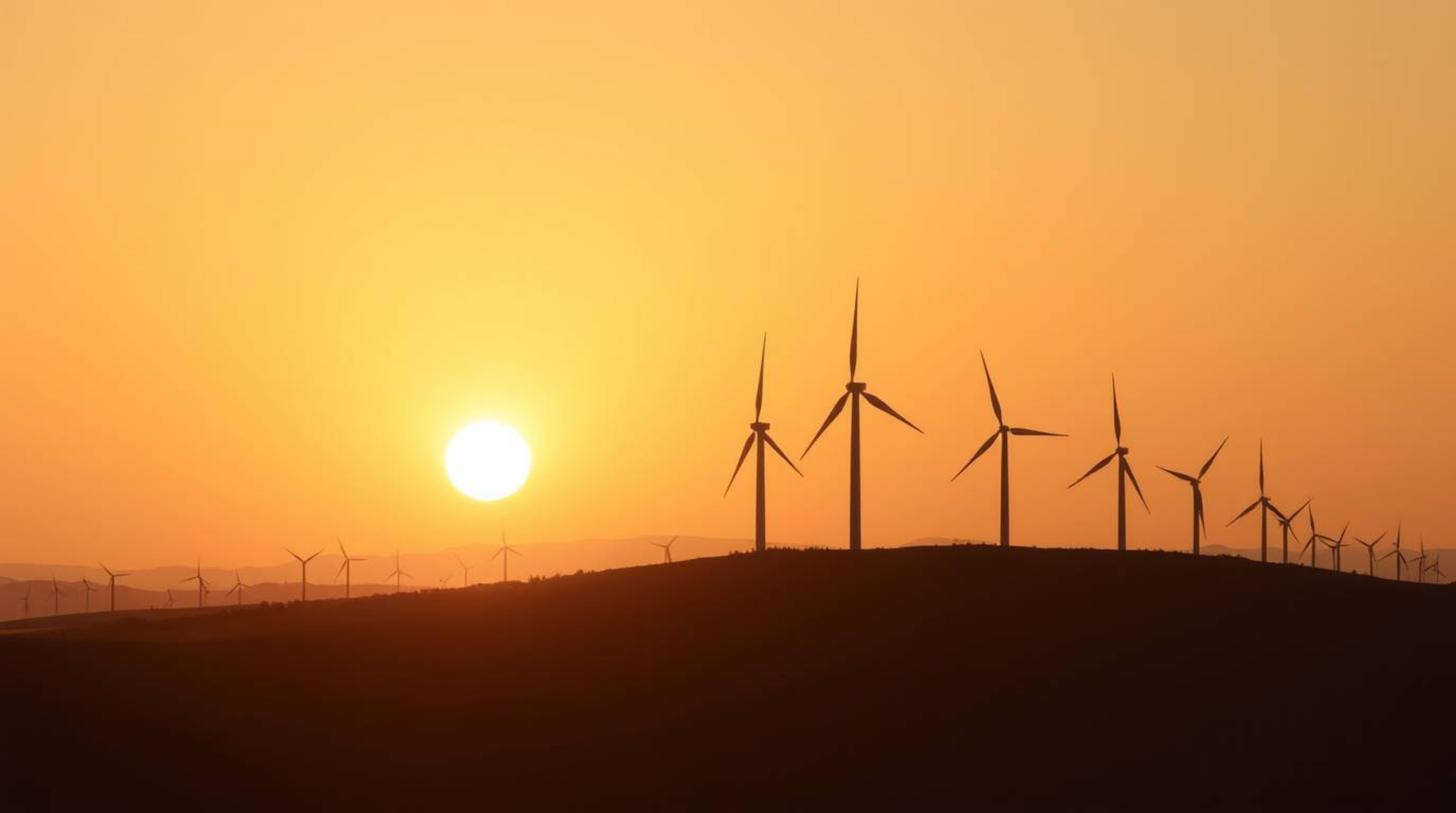 A captivating photograph showcases a serene sunset painting the sky behind a line of wind turbines perched atop a gentle hill