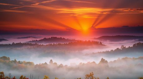 A captivating landscape painting depicts fog-shrouded hills bathed in the warm glow of a setting sun.  Bright lens flares and