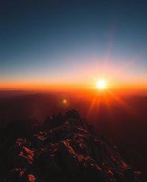 This is a high-contrast image of a sunset as viewed from a high alpine peak. The scene is characterized by strong, hard light