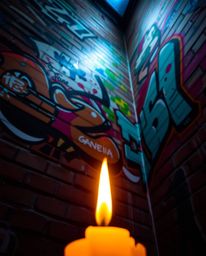Warm candlelight illuminates colorful graffiti splashes on a brick wall, creating a dramatic upward view of the street art ae