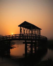An old wooden bridge crosses over a river during a vibrant sunset. The scene is enveloped in a soft, cinematic haze with a ge
