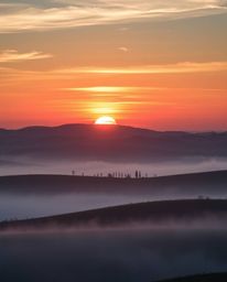 A photograph of a sunset over misty rolling hills in Tuscany, showcasing iridescent colors and their reflections on the lands