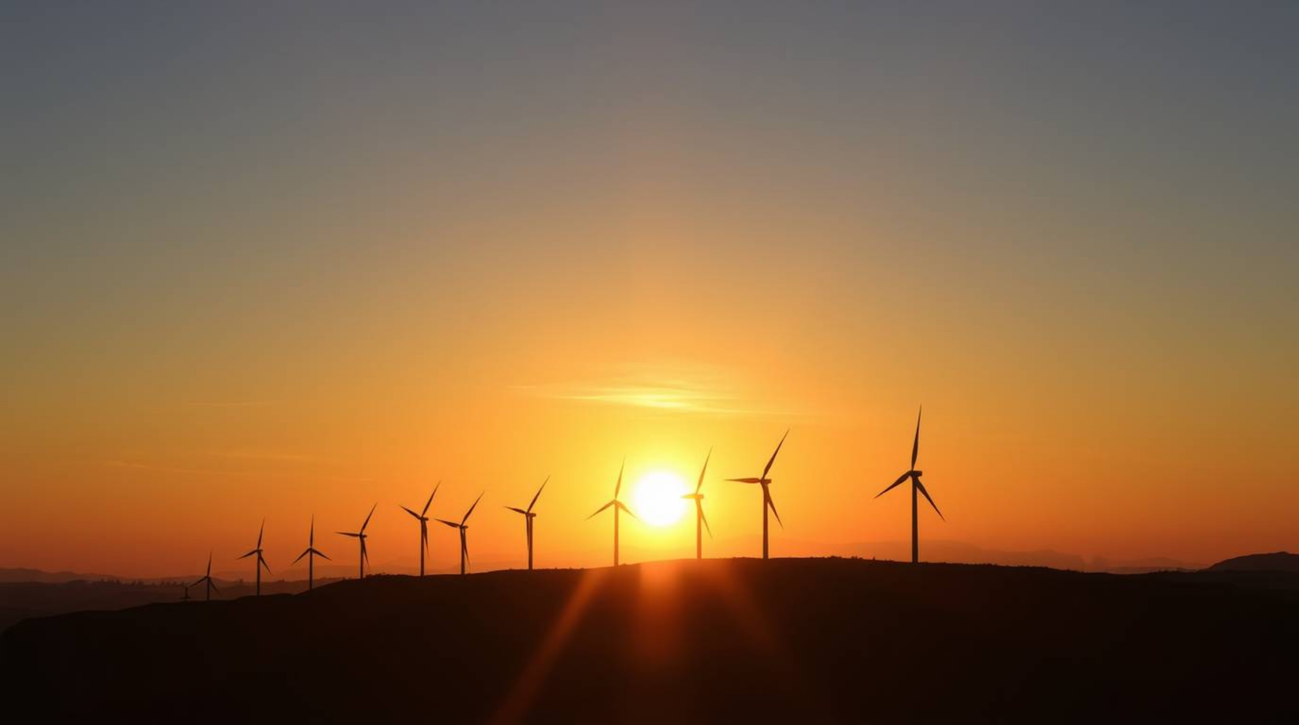 A captivating image showcases a warm, golden sunset radiating behind a picturesque row of wind turbines perched atop a hill. 