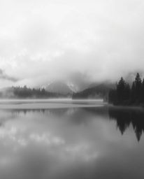 A classic black and white photograph depicts the reflections of fiery clouds in a foggy mountain lake. Characterized by a tim