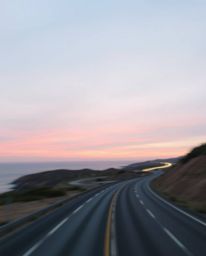 This long exposure photograph captures a soft pastel sunset sky over a winding coastal highway, creating a gentle and soothin