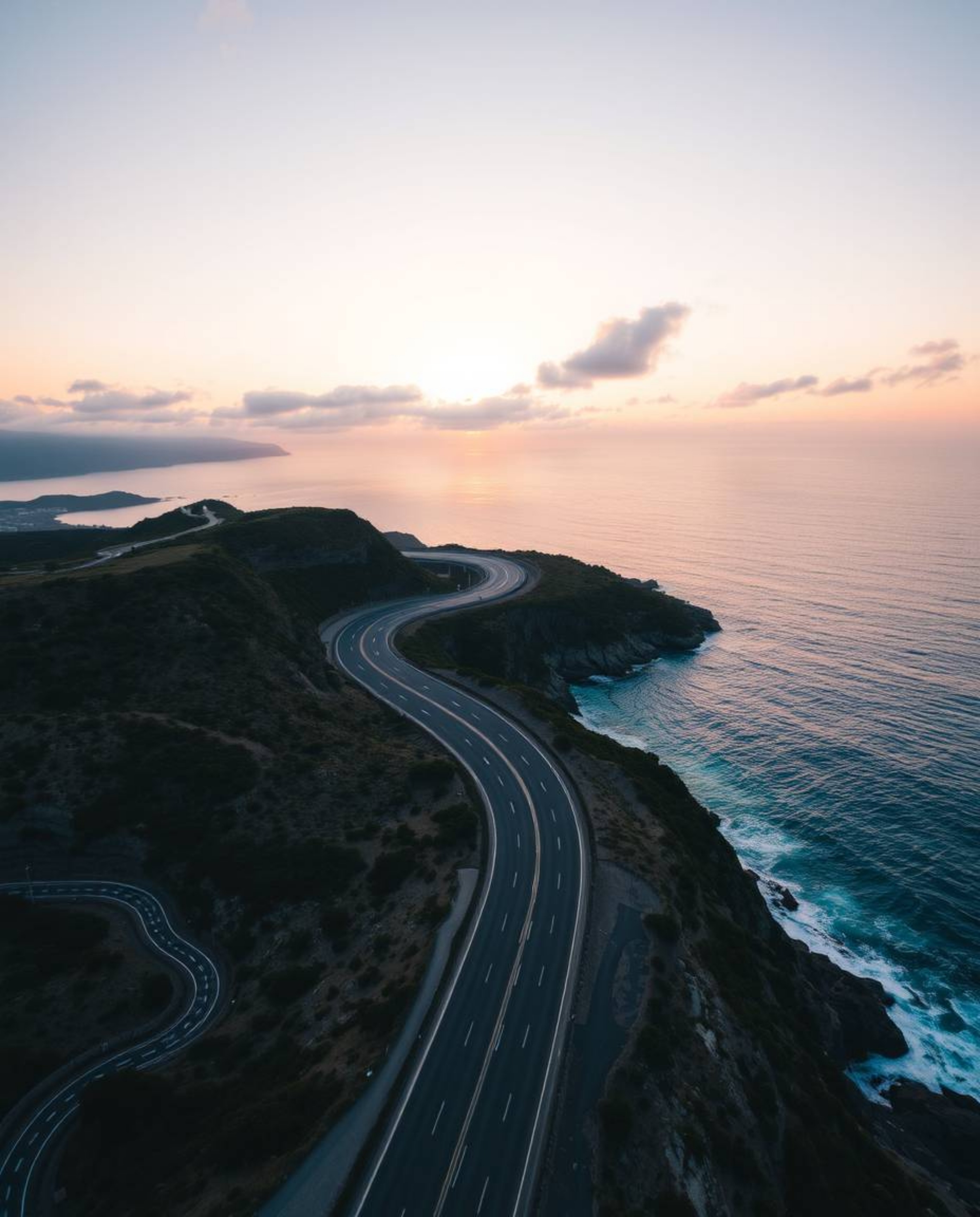 This is a high-angle perspective view looking down on a winding coastal highway beneath a beautiful sunset sky. This elevated
