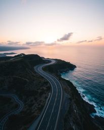 This is a high-angle perspective view looking down on a winding coastal highway beneath a beautiful sunset sky. This elevated