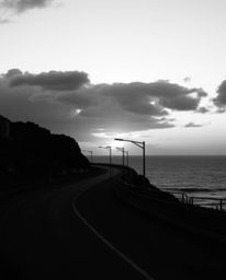 This is a candid black and white street photograph of a sunset sky over a winding coastal highway. With its timeless and eleg