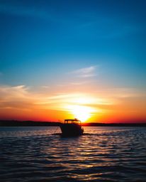 A silhouetted boat is seen drifting on a lake at sunset, created by a strong backlighting effect. The color palette is domina