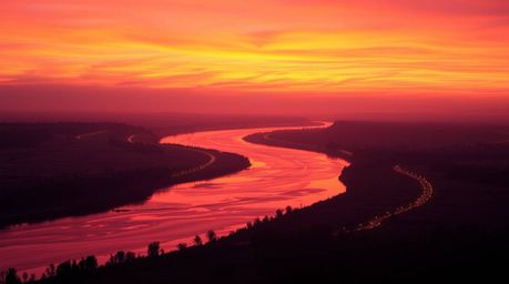 A captivating image of a winding river bathed in the warm glow of candlelight beneath a dramatic orange-pink sunset sky.  The