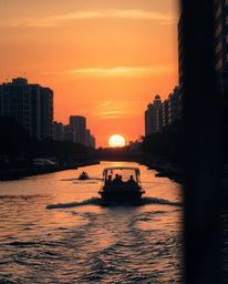 Candid street photography from a low angle, a boat drifts on a sunset lake, warm reds, oranges and yellows dominate the urban