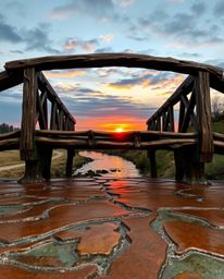 This is a bronze sculpture of an old wooden bridge over a river at sunset, crafted on a cast bronze surface. The piece is def