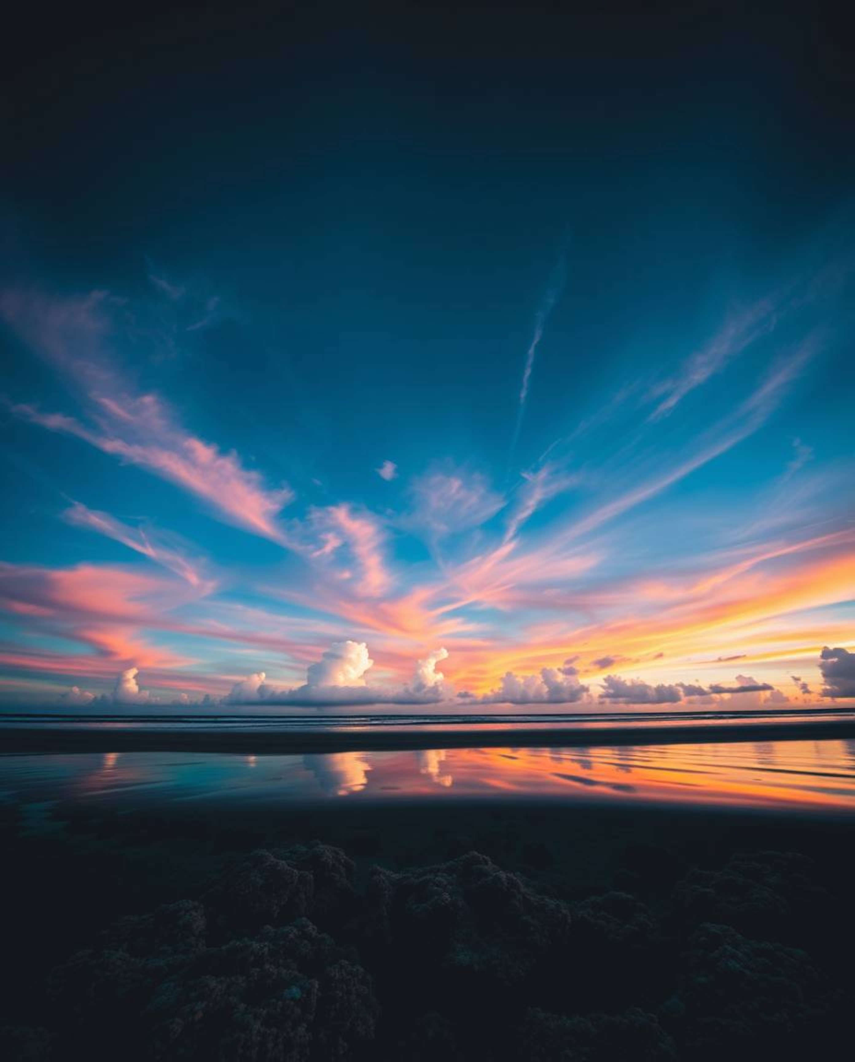 A striking high-angle perspective captures a colorful sky over a coral reef at low tide. The image employs high-contrast and 