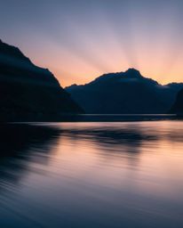 A stunning long exposure photograph of a glassy fjord reflecting a sunset, enhanced by dramatic volumetric lighting. This ima